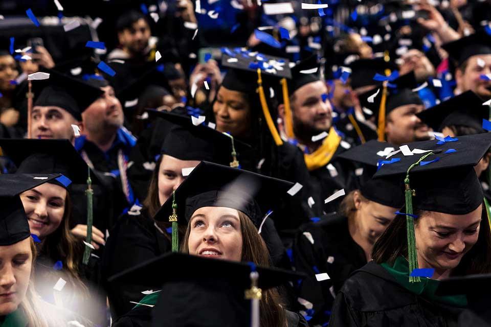 A student in cap and gown surrounded by a crowd of graduates looks up as confetti falls