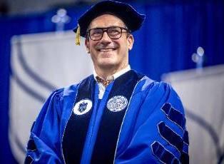 Actor Jon Hamm wearing a graduation cap and gown smiles while standing in front of 博彩网址大全 logos on a stage.